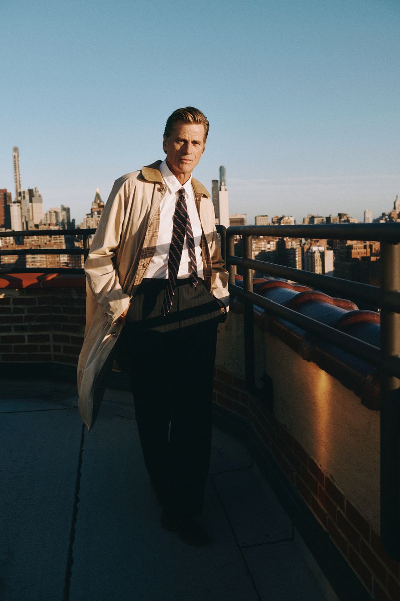 Adult male in a tan trench coat stands on a brick rooftop with a partial railing, set against a cityscape and blue sky background.