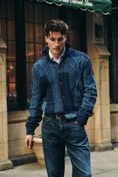 Man wearing a blue cable-knit cardigan and dark jeans standing outdoors in front of a stone building facade