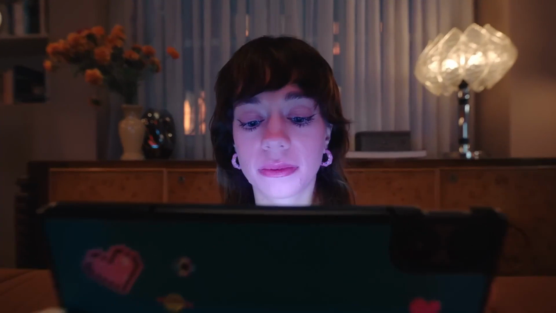 Woman looking at digital tablet in indoor setting with a decorative lamp, dresser, and flower vase in the background at nighttime.