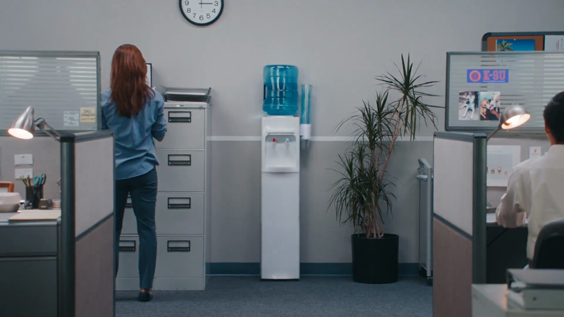 Office interior with a woman at a filing cabinet, a water cooler, and a man at a desk with partitions, providing a workspace scene.