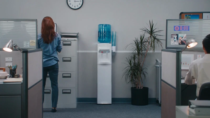 Office interior with a woman at a filing cabinet, a water cooler, and a man at a desk with partitions, providing a workspace scene.