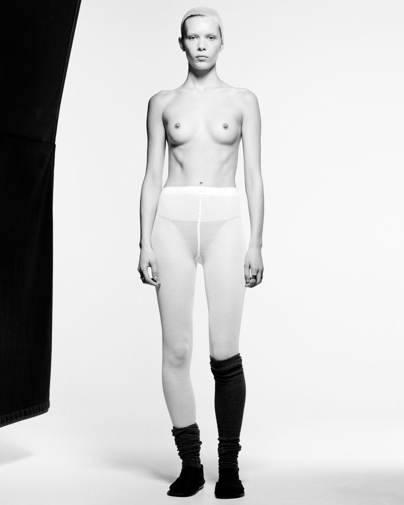 Full-length studio shot of a slender adult female with cropped hair, pale skin, wearing semi-sheer tights and black socks against a white background.