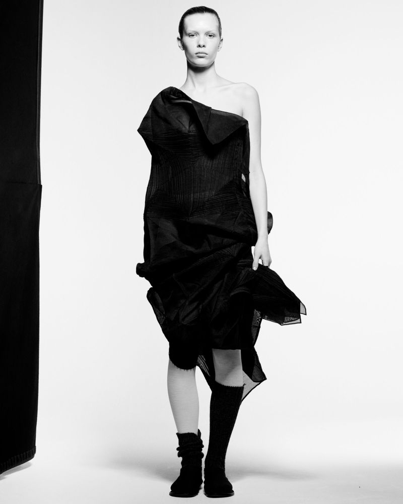 Monochrome fashion shot: Woman poses in avant-garde black dress with knee-high socks and boots against a stark white backdrop.