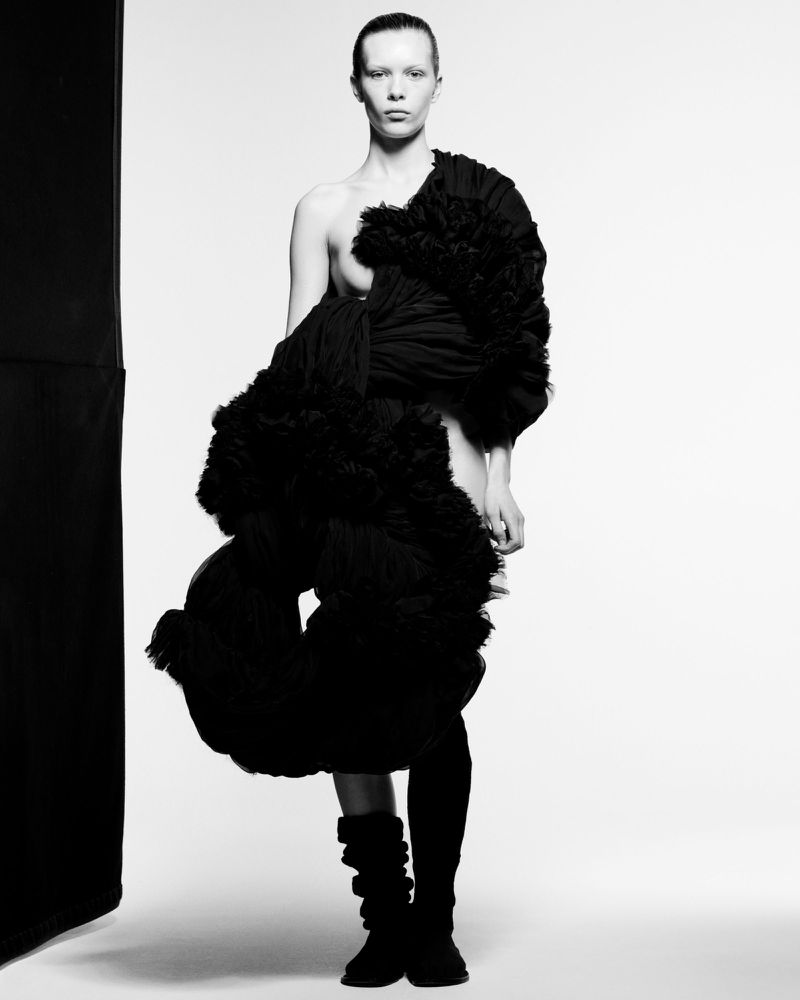 Monochrome studio shot of a slender adult female model in dark hosiery and sculptural black tulle garment, posing against a white backdrop.