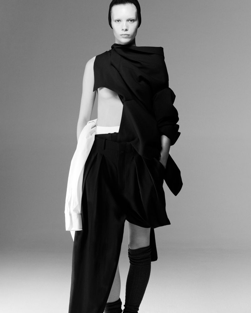 Monochrome fashion shot: A slender, pale-skinned model poses in a striking black and white outfit, knee socks, against a gray backdrop.
