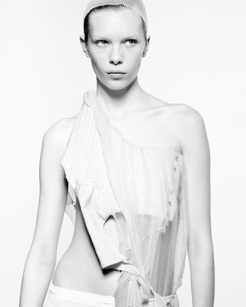 Monochrome portrait of a fair-skinned woman wearing a sheer head covering and asymmetrical light-colored garment against a white backdrop.