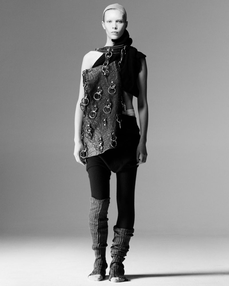 Monochrome studio shot of a person with a shaved head wearing a dark, textured garment with ring hardware, leg warmers, and black leggings.