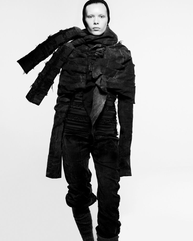 A monochrome full shot captures a slim person with light skin in elaborate dark, textured clothing and a scarf, against a stark white background.