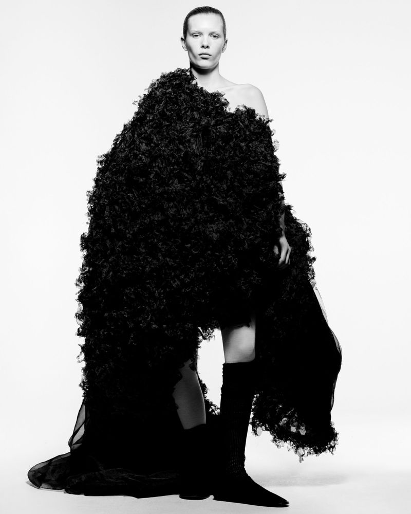 Black-and-white fashion portrait of a slender adult woman wearing oversized, textured coat and knee-high boots against white backdrop.
