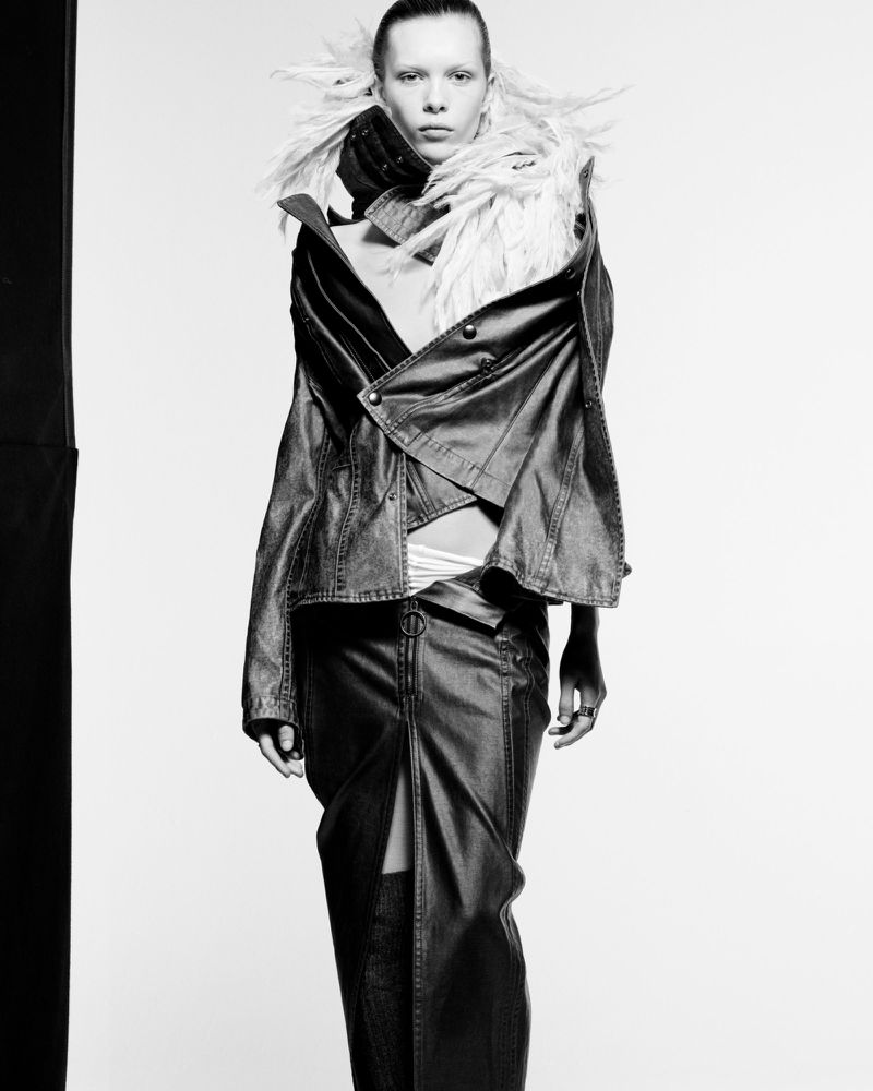 Black and white fashion shot of a woman in a dark jacket and skirt with a feathered collar against a plain background in a studio.