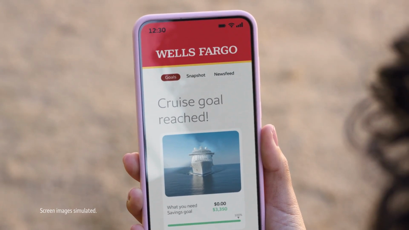 Close-up of a person holding a smartphone showing the Wells Fargo app with a cruise ship image and text indicating a goal has been reached.