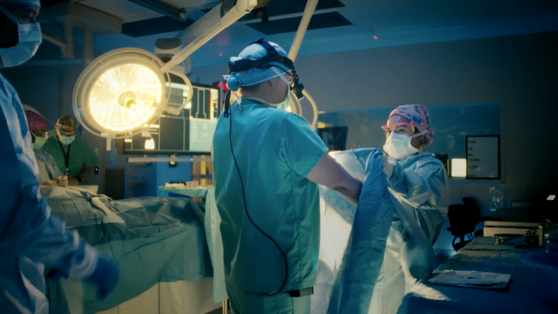 Operating room scene: surgeons in scrubs and masks perform surgery under bright lights, showcasing medical procedure in a clinical setting.
