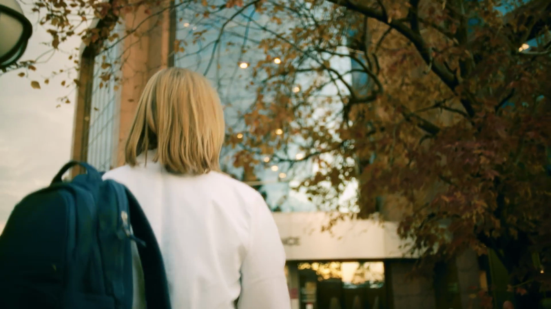 Woman with backpack walking towards a building framed by trees. Outdoor shot with natural light and foliage.