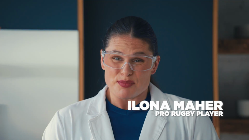 Portrait of woman wearing lab coat and safety glasses in a brightly lit indoor environment, with visible text identifying her as Ilona Maher.
