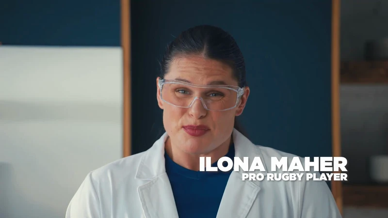 Portrait of woman wearing lab coat and safety glasses in a brightly lit indoor environment, with visible text identifying her as Ilona Maher.