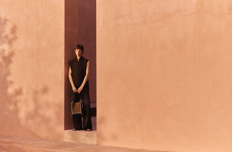 A slender woman in a black sleeveless outfit poses in a peach-colored architectural alcove, holding a black and tan handbag.