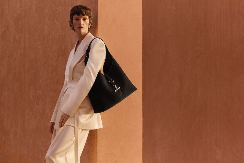Woman in cream suit with black handbag posing against orange wall in studio setting, fashion editorial, neutral color palette.