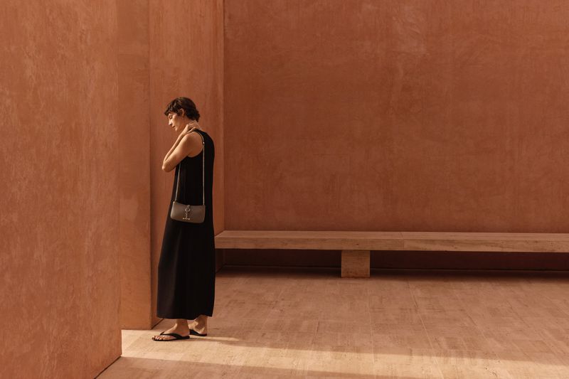 A woman wearing a long black dress and purse stands in front of a textured reddish-brown wall in a simple, naturally lit indoor space.