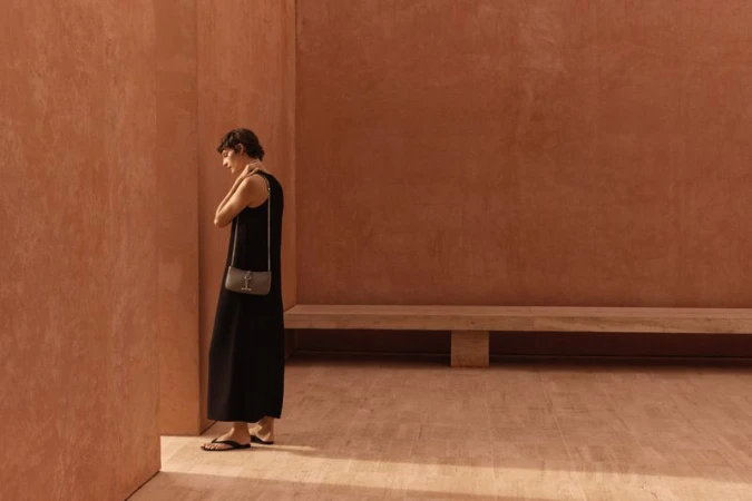 A woman wearing a long black dress and purse stands in front of a textured reddish-brown wall in a simple, naturally lit indoor space.