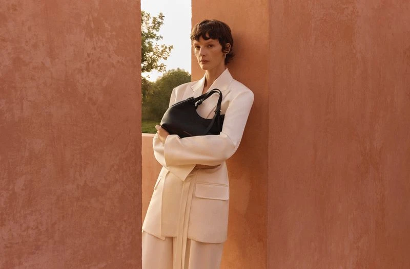 Elegant woman in a cream-colored suit leans against a textured wall while holding a black handbag in a fashionable outdoor setting.