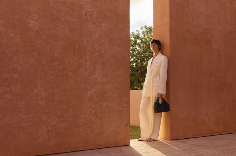 A woman in a white suit poses between terracotta walls outdoors, holding a dark handbag in a fashion-style editorial shot.