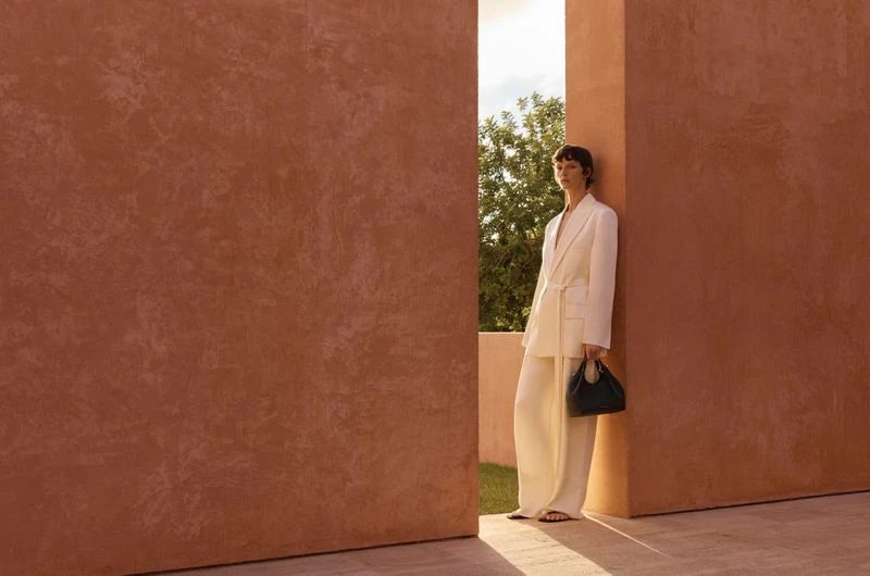 A woman in a white suit poses between terracotta walls outdoors, holding a dark handbag in a fashion-style editorial shot.