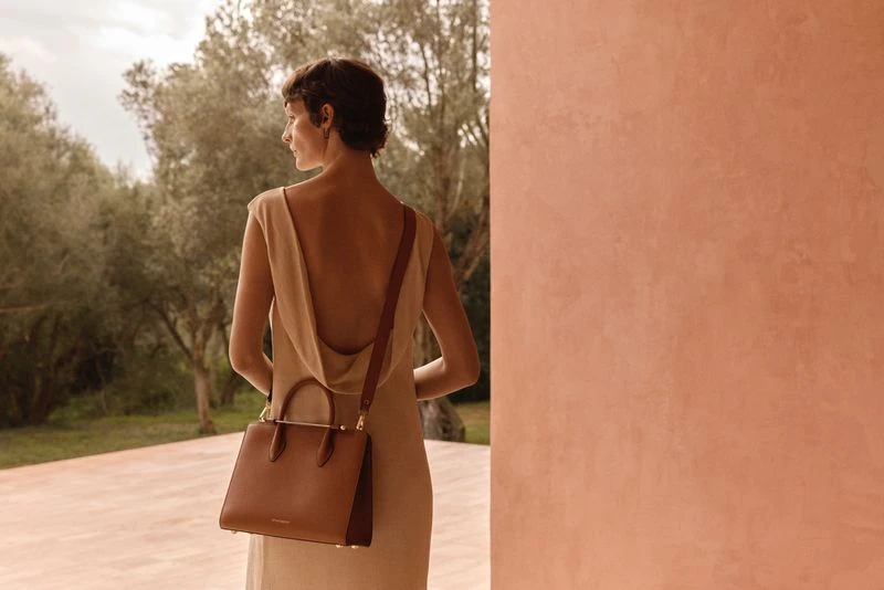 Adult woman in beige dress with brown leather purse outdoors against terracotta wall and green trees in a fashion editorial.