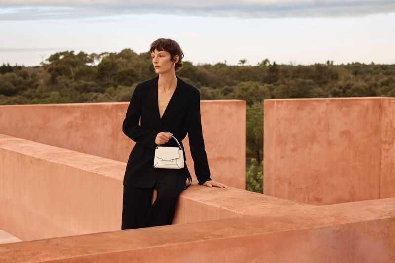 Adult female in black suit poses outdoors with small white handbag near peach-colored wall, trees and cloudy sky visible in background.