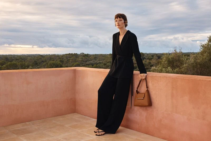 An adult woman in a black pantsuit poses against a terracotta balcony railing, holding a brown leather handbag with trees and cloudy sky.