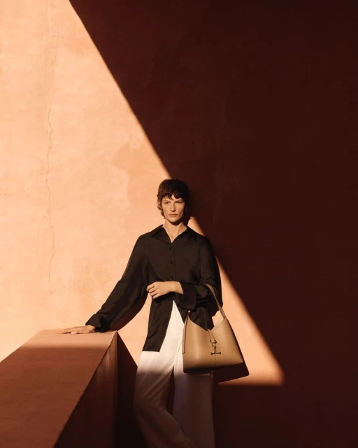 Woman poses in black top and white pants with tan purse against a textured wall in sunlight, fashion editorial style.
