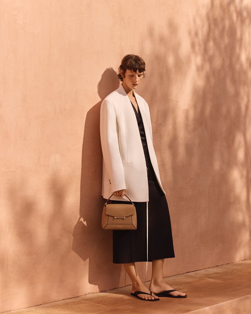Woman in a white jacket and black skirt holds a handbag against a textured peach wall, casting soft shadows in a fashion editorial.