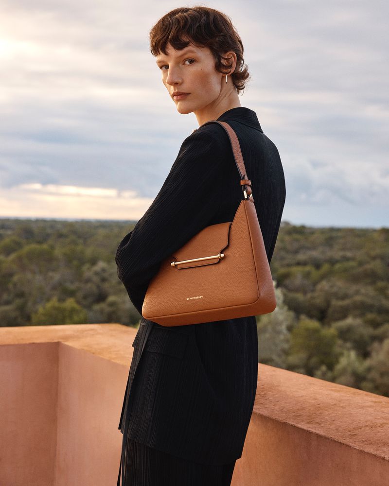 Adult woman poses with a brown leather handbag on a rooftop overlooking a green tree canopy under a cloudy sky during the daytime.