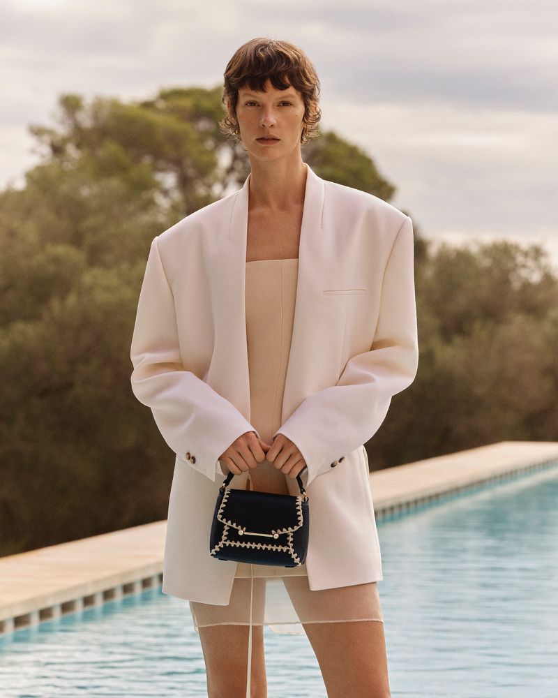 Adult woman in a white blazer and tan dress holding a dark handbag outdoors near the edge of a pool with trees in the background.