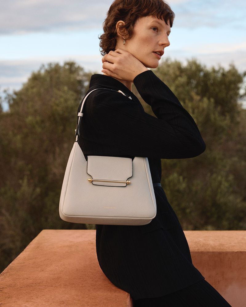 A woman wearing a black blazer poses outdoors with a white leather purse in a fashion editorial shot.