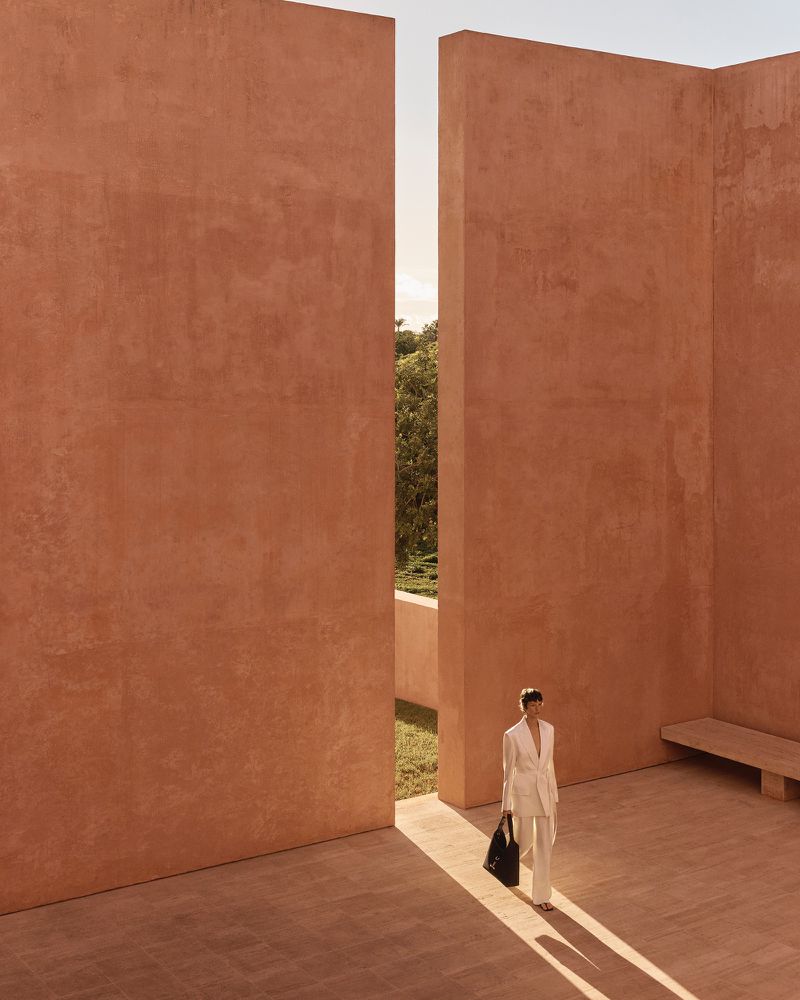 Woman in a light suit walks through a terracotta-walled courtyard with dappled sunlight and minimalistic architecture and a black tote bag.