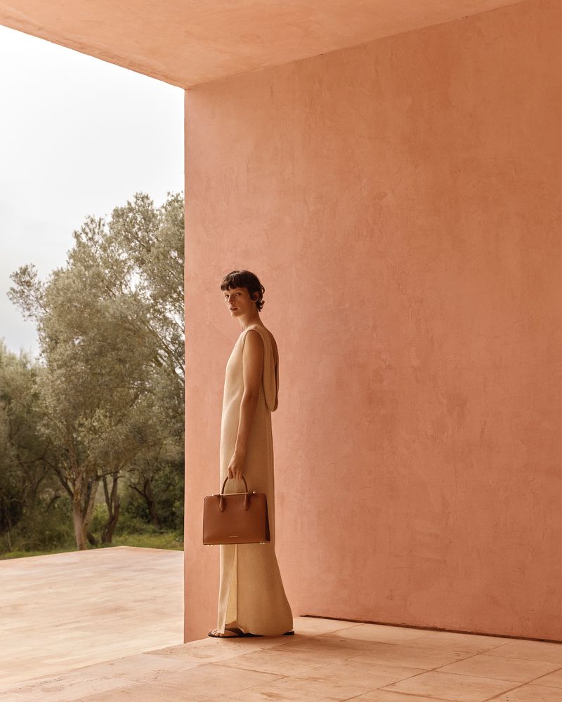 Woman in neutral dress stands near salmon wall holding handbag, bathed in soft light. Outdoor setting with green trees in the distance.