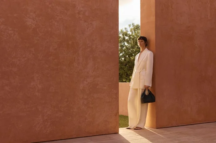 Model in white suit holding a black handbag leaning against a textured terracotta wall outdoors