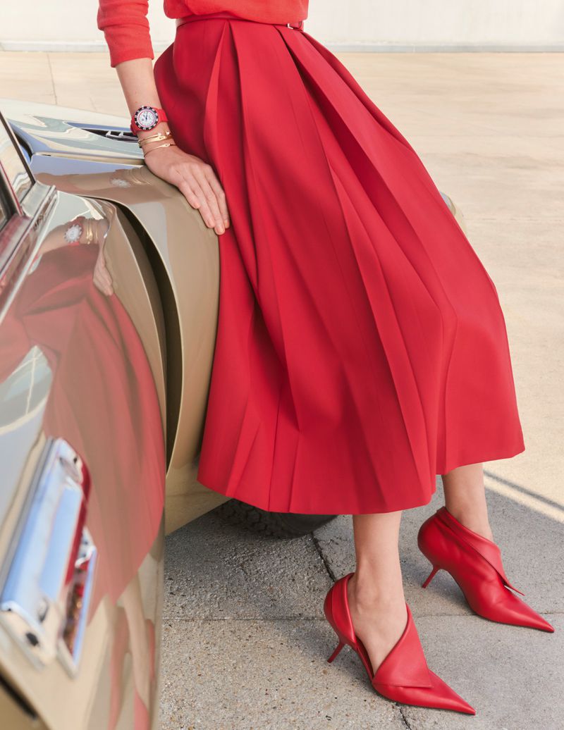 Woman in a red sweater and skirt posing with a tan car, wearing matching red heels and jewelry, outdoors.