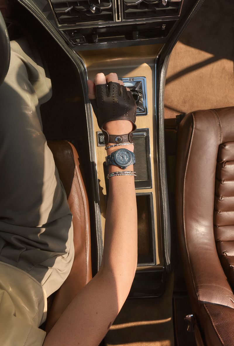 Interior car view from above with a person's arm on the console wearing a watch, gloves, and bracelets near the gear stick.
