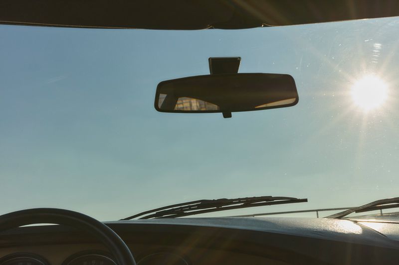 Interior view of a car showing the dashboard, steering wheel, rearview mirror, and windshield with the sun shining in a blue sky.