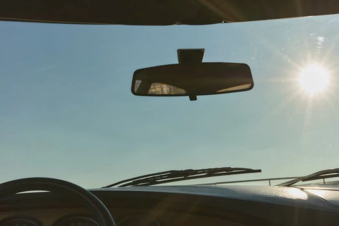 Interior view of a car showing the dashboard, steering wheel, rearview mirror, and windshield with the sun shining in a blue sky.