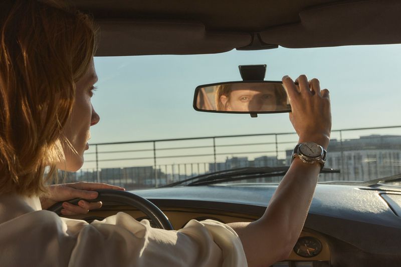 Woman driving a car, adjusting the rearview mirror with a city skyline visible in the background on a sunny day.