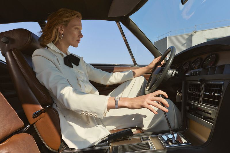 Adult woman in a white suit driving a classic brown interior car on a bright day with buildings visible in the background.