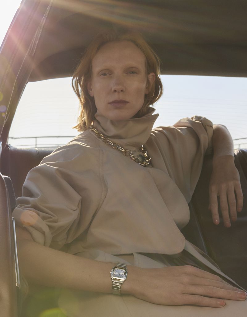 A woman with red hair sits inside the front seat of a car, wearing a beige blouse, silver watch, and a chunky chain necklace.