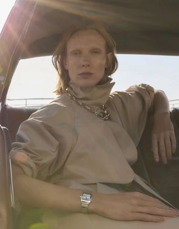 A woman with red hair sits inside the front seat of a car, wearing a beige blouse, silver watch, and a chunky chain necklace.