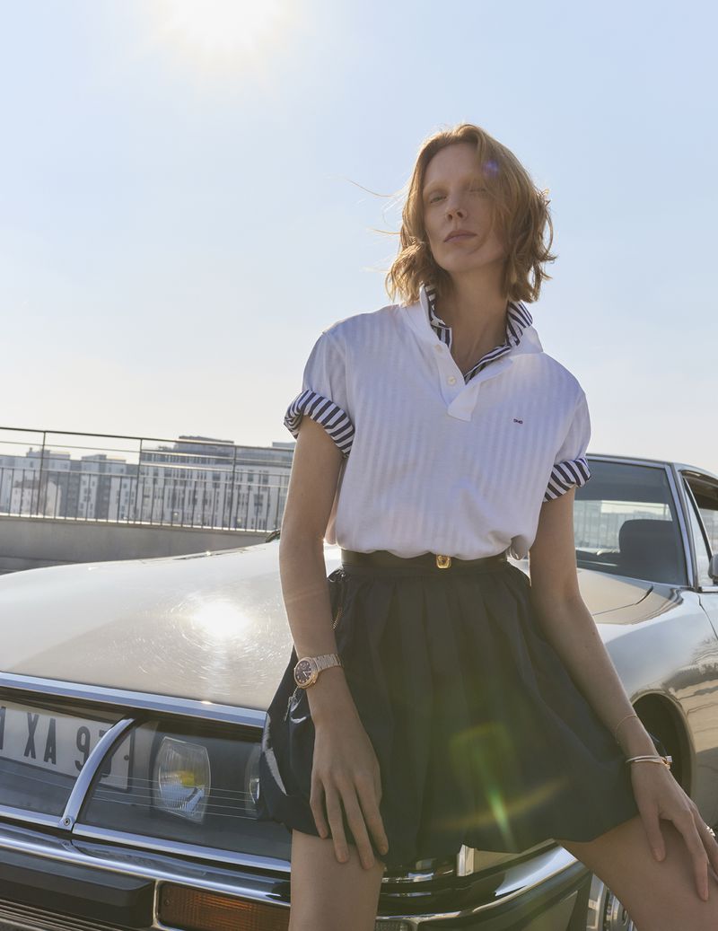 Fashion shot of woman in polo and skirt leaning on vintage car under bright sky with city buildings in distance, showcasing lifestyle.