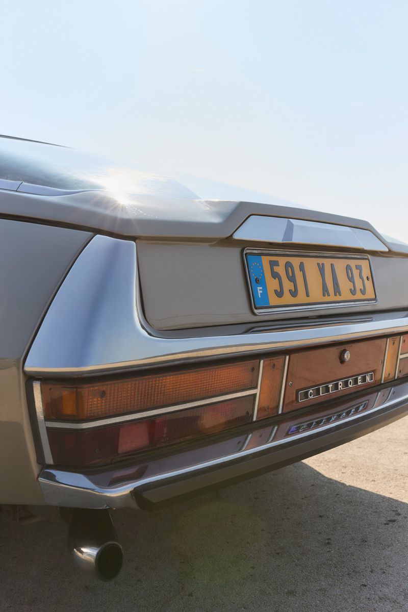 Rear view of a tan 1970s Citroen SM with European license plate against a clear sky, showing the tail lights and exhaust pipe.