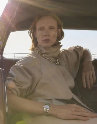 Model Annely Bouma seated in a vintage car wearing a beige coat and chunky gold chain necklace for Madame Figaro