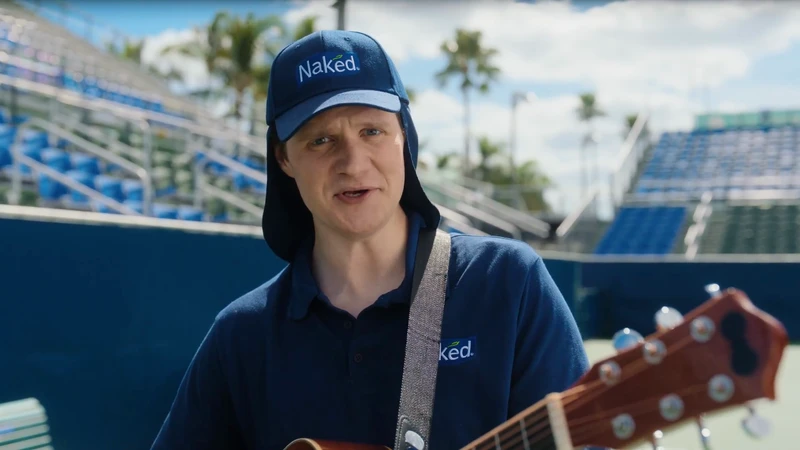 Adult male wearing a blue polo shirt and cap, playing guitar on an outdoor tennis court; the man is the main focus of the shot.