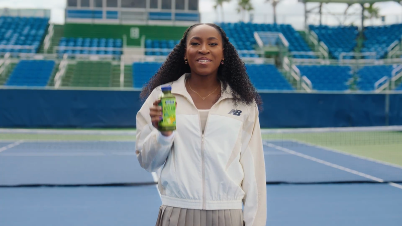 Adult woman in a sports jacket poses on a tennis court holding a green drink, with stadium seating visible in the background.